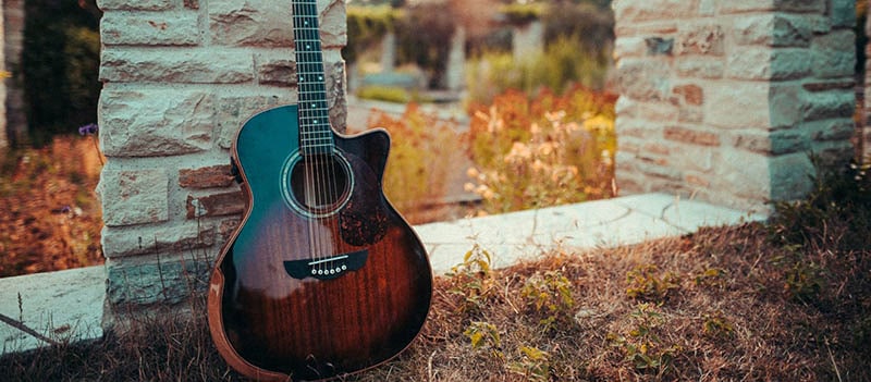 A Harley Benton CLC-650SM-CE VS Solid Wood acoustic guitar leaning against a brick pillar