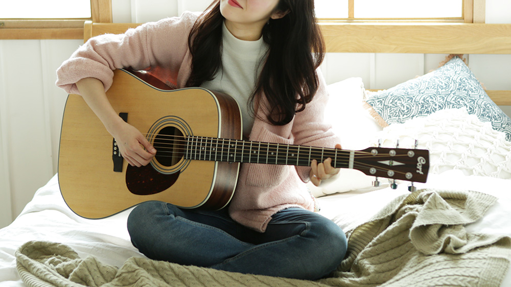 A Cort acoustic guitar being played by a woman in a pink top and jeans sitting cross-legged on her bed