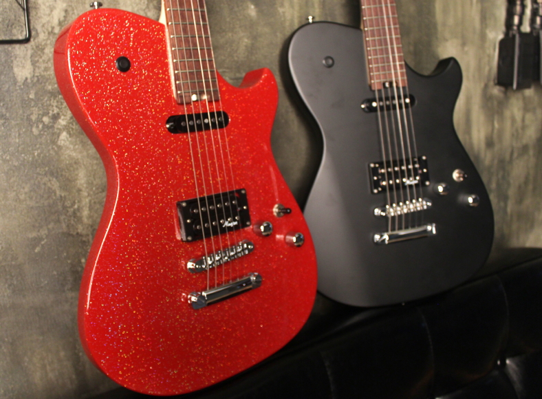 Two Cort MBC-1 guitars leaning against a wall; one in red sparkle finish on the left, and black finish on the right