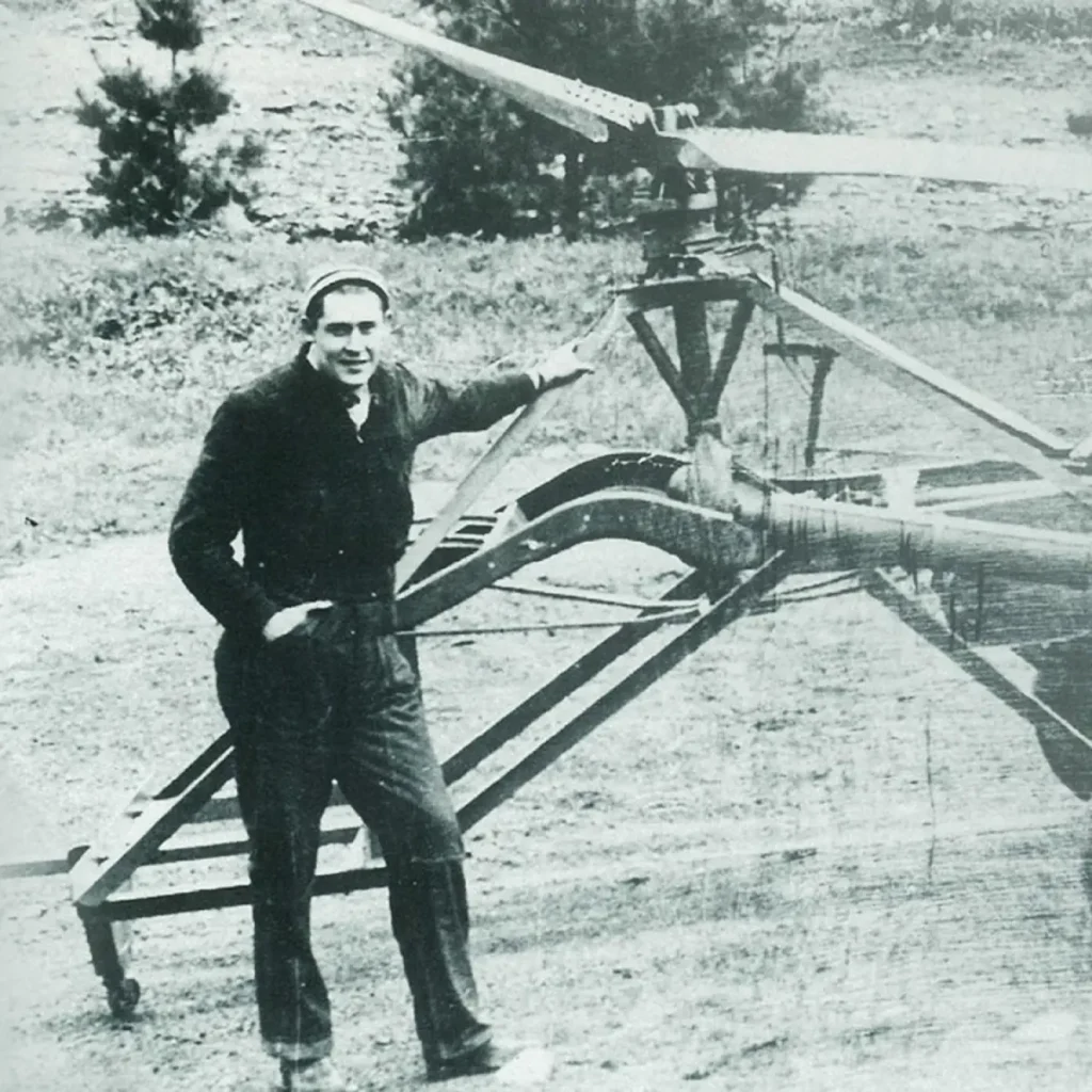 The image is a black-and-white photograph featuring Charles Kaman, the founder of Ovation Guitars. He is standing outdoors, wearing a dark jacket and trousers with a cap. Kaman appears relaxed, with one hand resting on a piece of machinery with rotor blades on top and the other hand in his pocket. The setting is an open area with trees and a dirt ground in the background, giving the image a pioneering, industrial feel. This photograph highlights Kaman’s background in aerospace engineering, which later influenced his innovations in guitar design.