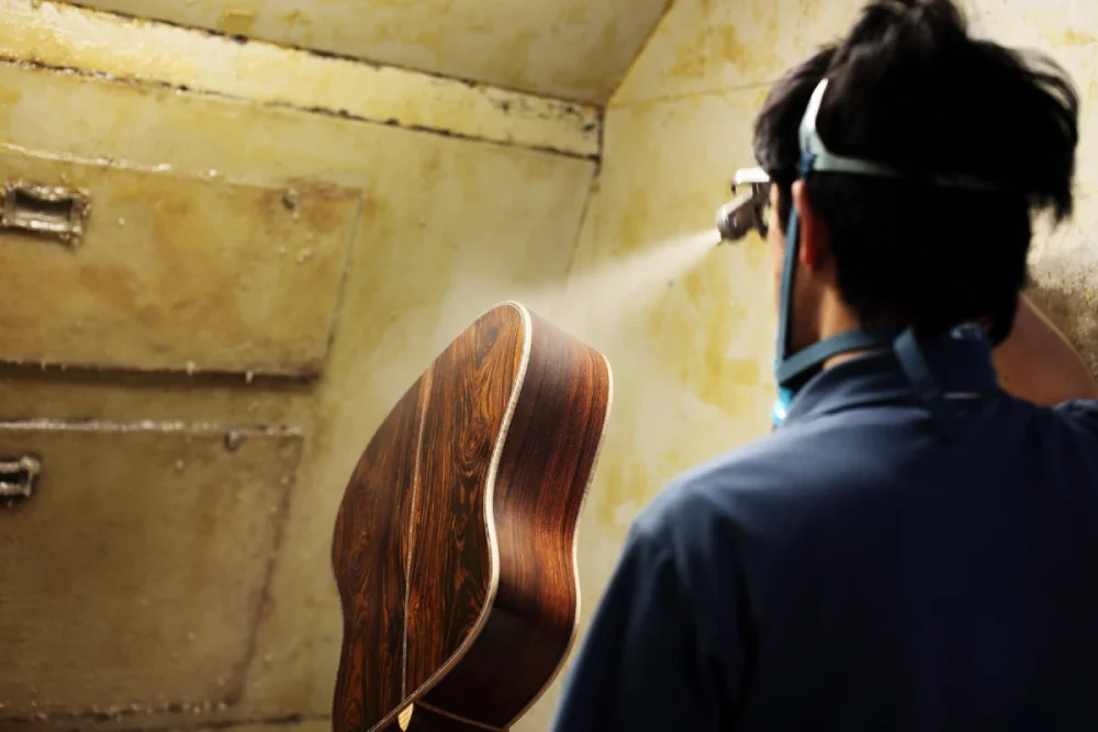 Worker at the Takamine factory spraying a glossy finish onto the back of an acoustic guitar, wearing protective eyewear and mask.