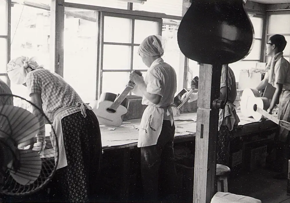 Vintage black-and-white photo of workers in a Takamine guitar workshop, wearing aprons and head coverings, handcrafting acoustic guitars by the window with unfinished instruments on the benches.
