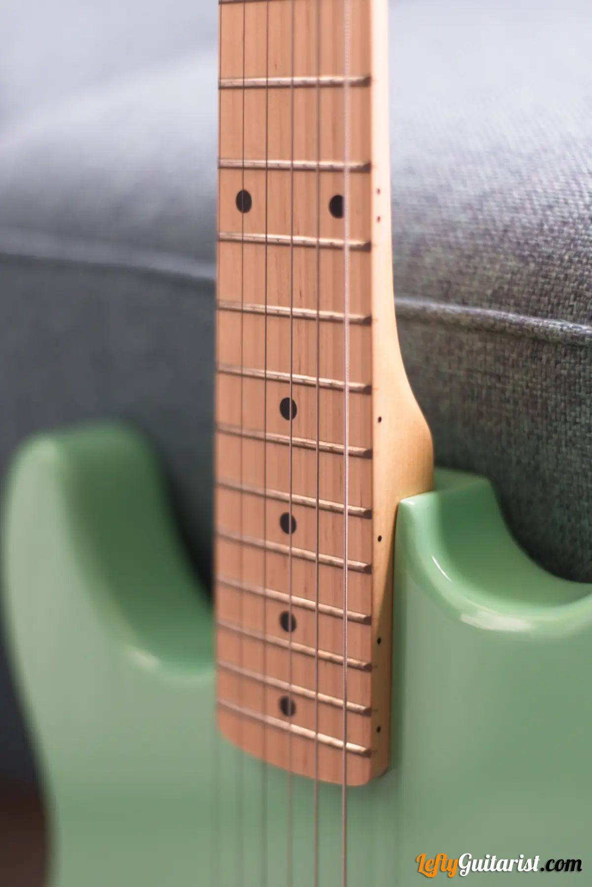 Photo of a left-handed Fazley Custom Series Hot Rod V2 Electric Guitar with Fixed Bridge in a Surf Green finish leaning against a blue sofa. Close-up of neck and body.