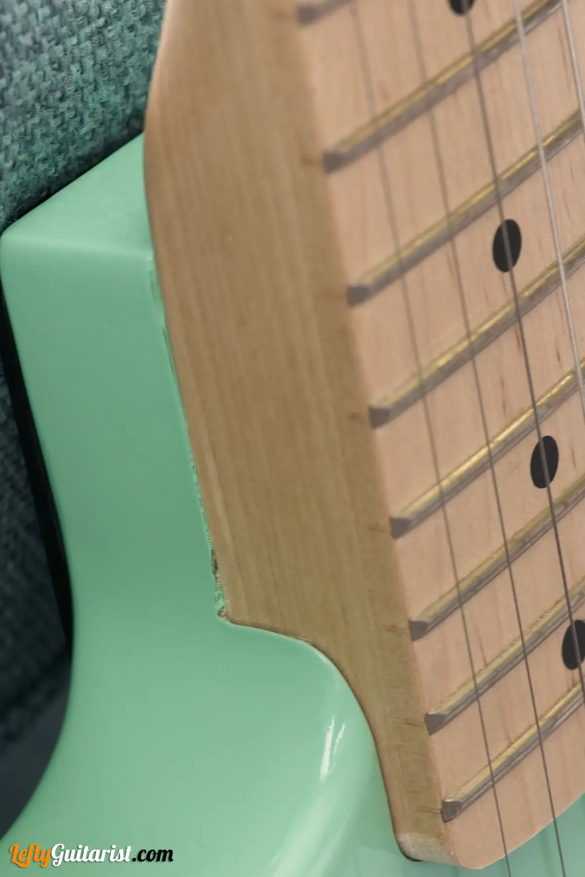 Photo of a left-handed Fazley Custom Series Hot Rod V2 Electric Guitar with Fixed Bridge in a Surf Green finish leaning against a blue sofa. Close-up of the neck pocket and a slightly rough paint/sand job.