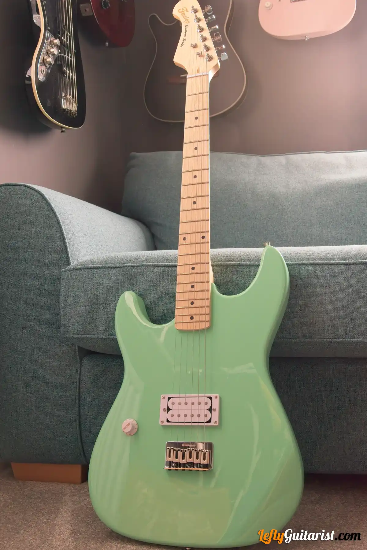 Photo of a left-handed Fazley Custom Series Hot Rod V2 Electric Guitar with Fixed Bridge in a Surf Green finish leaning against a blue sofa